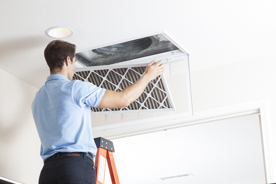 Technician cleaning a ceiling air duct filter while standing on a ladder in a home interior